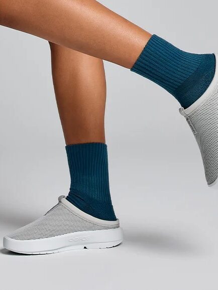 Person wearing blue socks and gray slip-on shoes raises one leg in a minimalist, gray background.