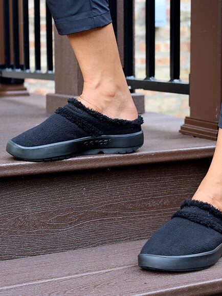 Person wearing black slip-on shoes with a fuzzy lining is standing on outdoor wooden steps near a black railing.