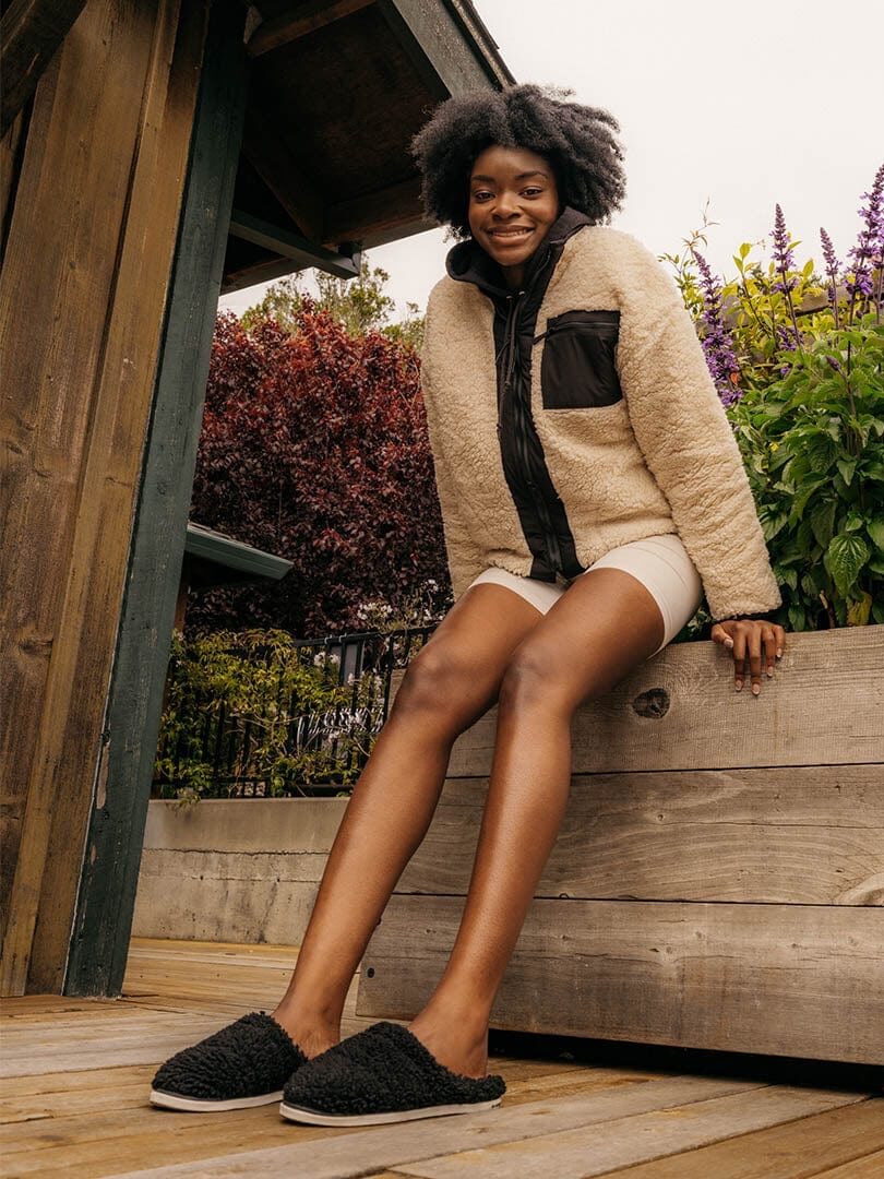 A person with curly hair sits on the edge of a wooden structure, wearing a beige and black jacket, shorts, and black slippers, with purple and green plants in the background.