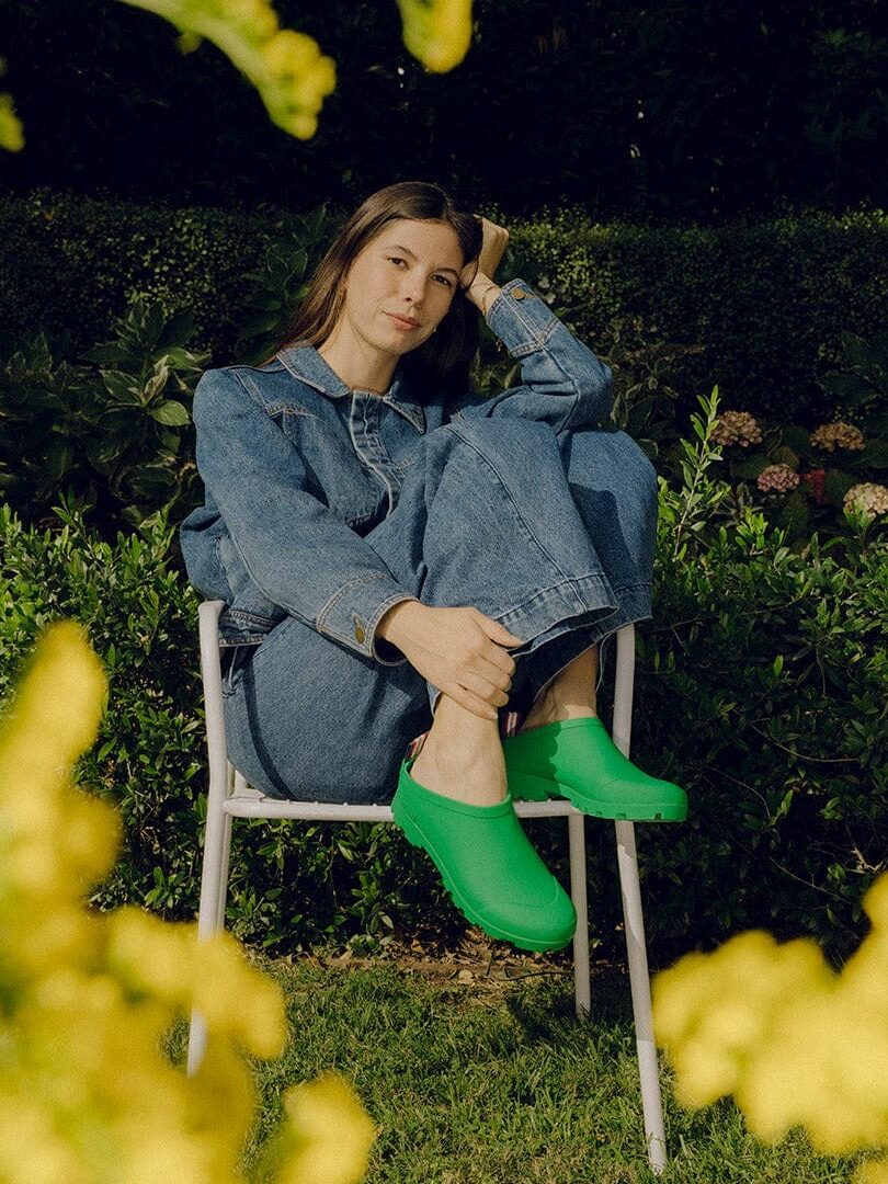 A person wearing a denim outfit and bright green shoes sits on a white chair in a garden, surrounded by lush greenery and yellow flowers.