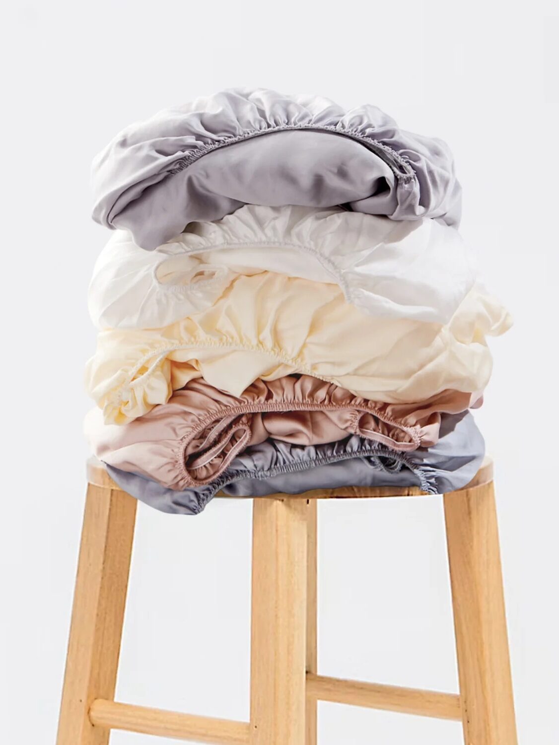 A stack of folded fitted sheets in various pastel colors is placed on a wooden stool against a plain white background.