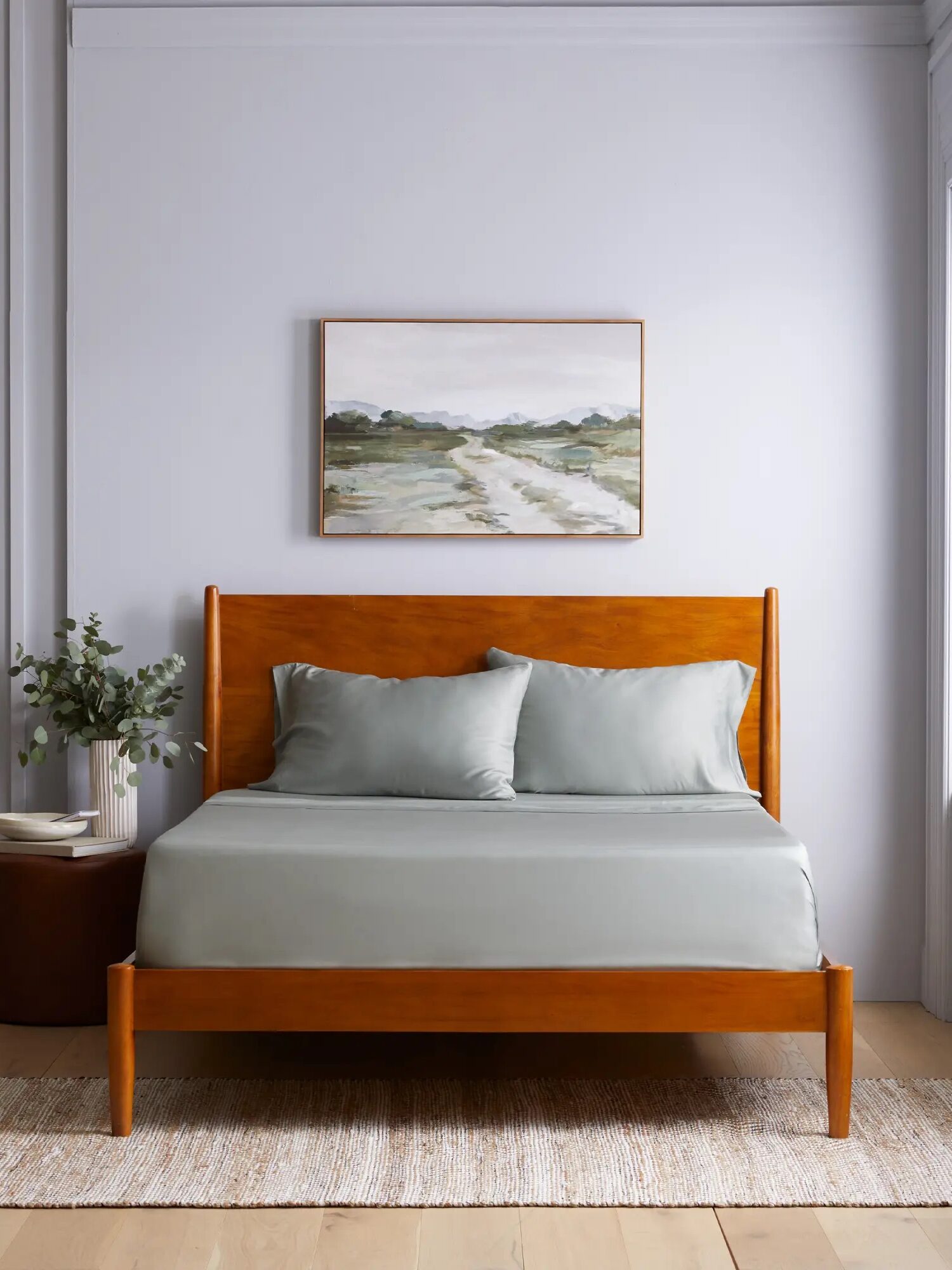 A neatly made bed with a wooden frame and light gray bedding is centered against a white wall. A landscape painting hangs above the bed, and a potted plant sits on a side table to the left.