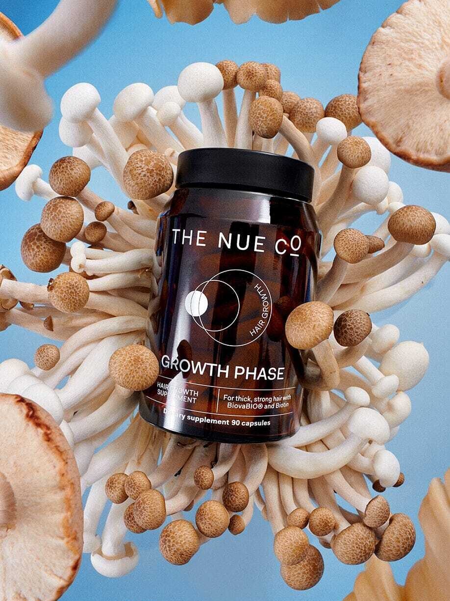 A bottle of 'The NUE Co' Growth Phase hair supplement surrounded by various types of mushrooms against a blue background.