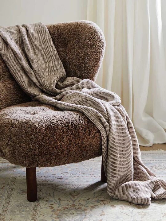 A brown, fluffy upholstered chair with wooden legs sits next to a window with white curtains. A beige, textured blanket is draped over the chair. .
