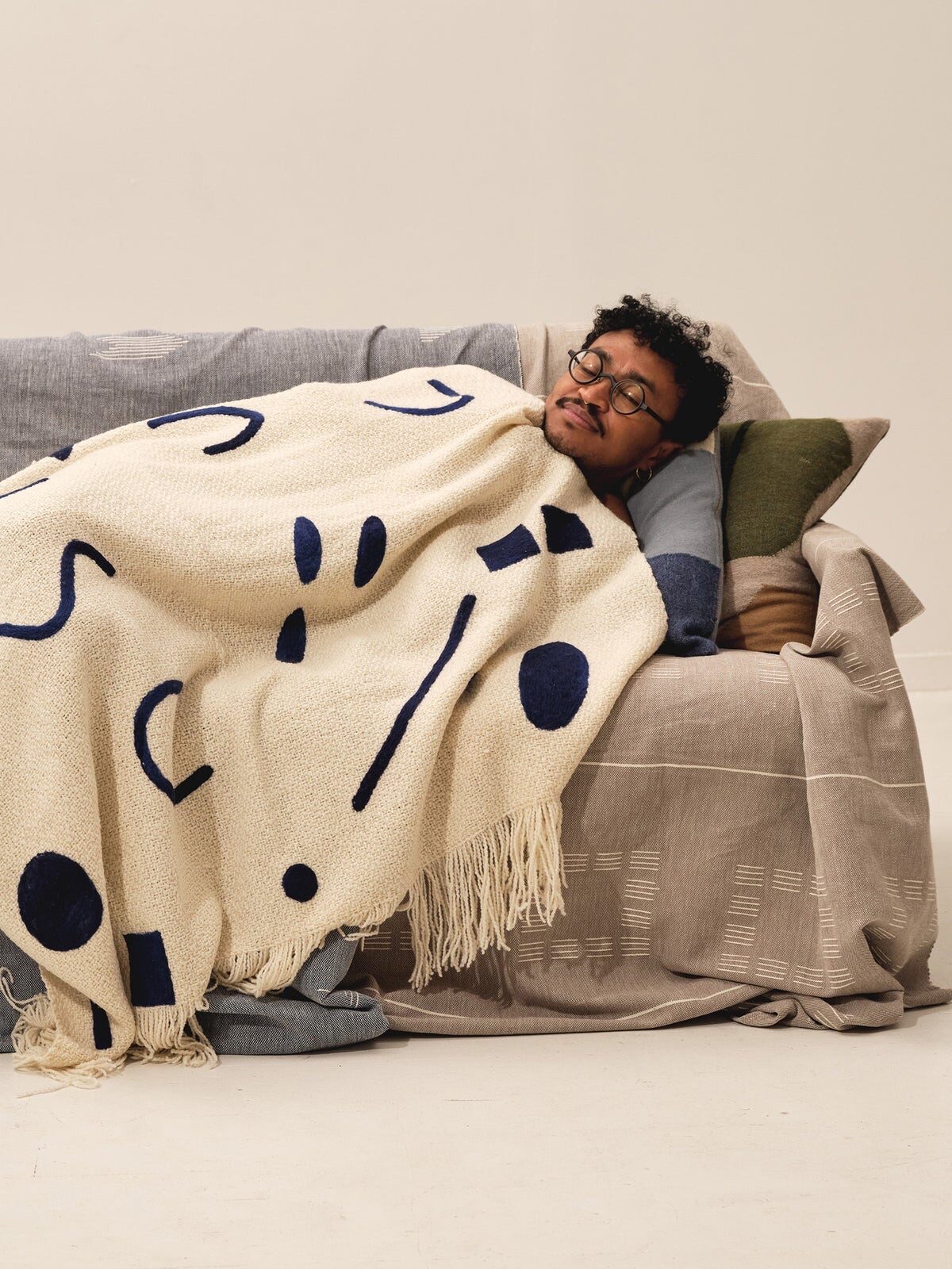 A person sleeps on a couch covered with blankets. They are wrapped in a beige blanket with abstract blue patterns.