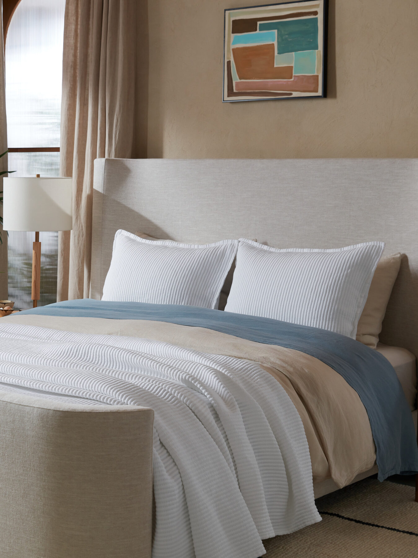A neatly made bed in a stylish bedroom with a large plant, two nightstands, and framed artwork on the beige wall. The bed features a beige headboard and neutral bedding with blue and white pillows.