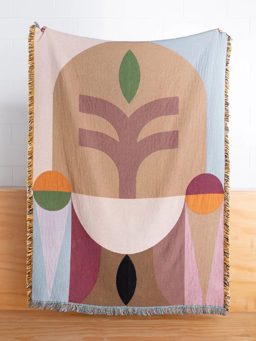 A woven tapestry with a geometric abstract design hangs against a wooden and white tiled wall. The design features shapes in beige, orange, green, pink, and black.