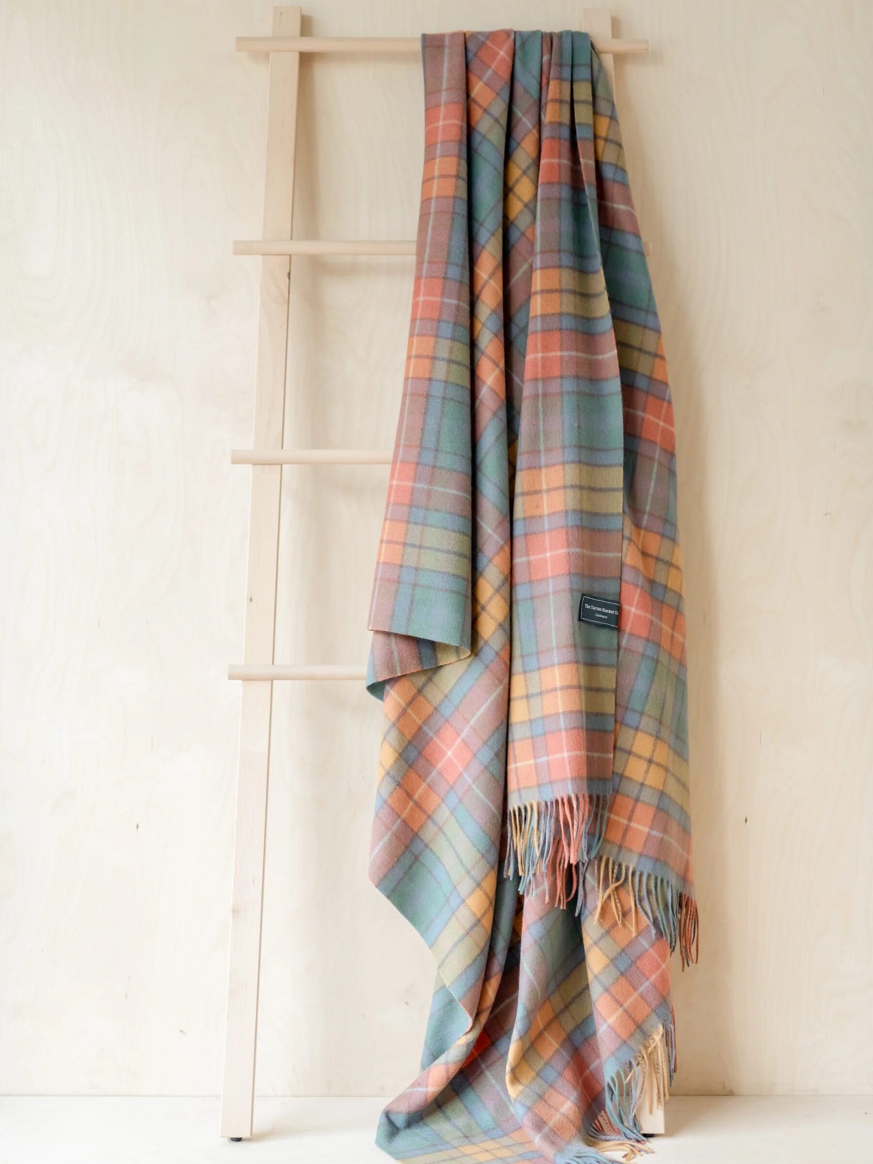 A multicolored plaid blanket is draped over a wooden ladder against a light-colored background.