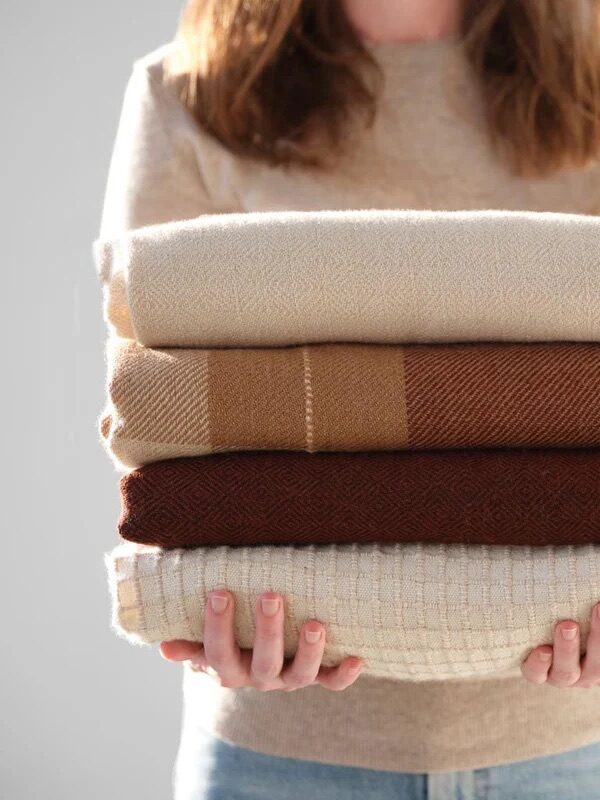 A person holds a stack of four folded sweaters in neutral colors, including white, beige, brown, and cream.