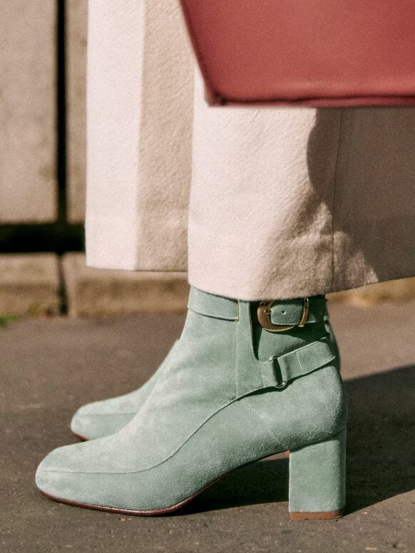 Close-up of a person standing on pavement wearing beige pants and mint green suede ankle boots with a buckle. A corner of a red handbag is visible above the pants.