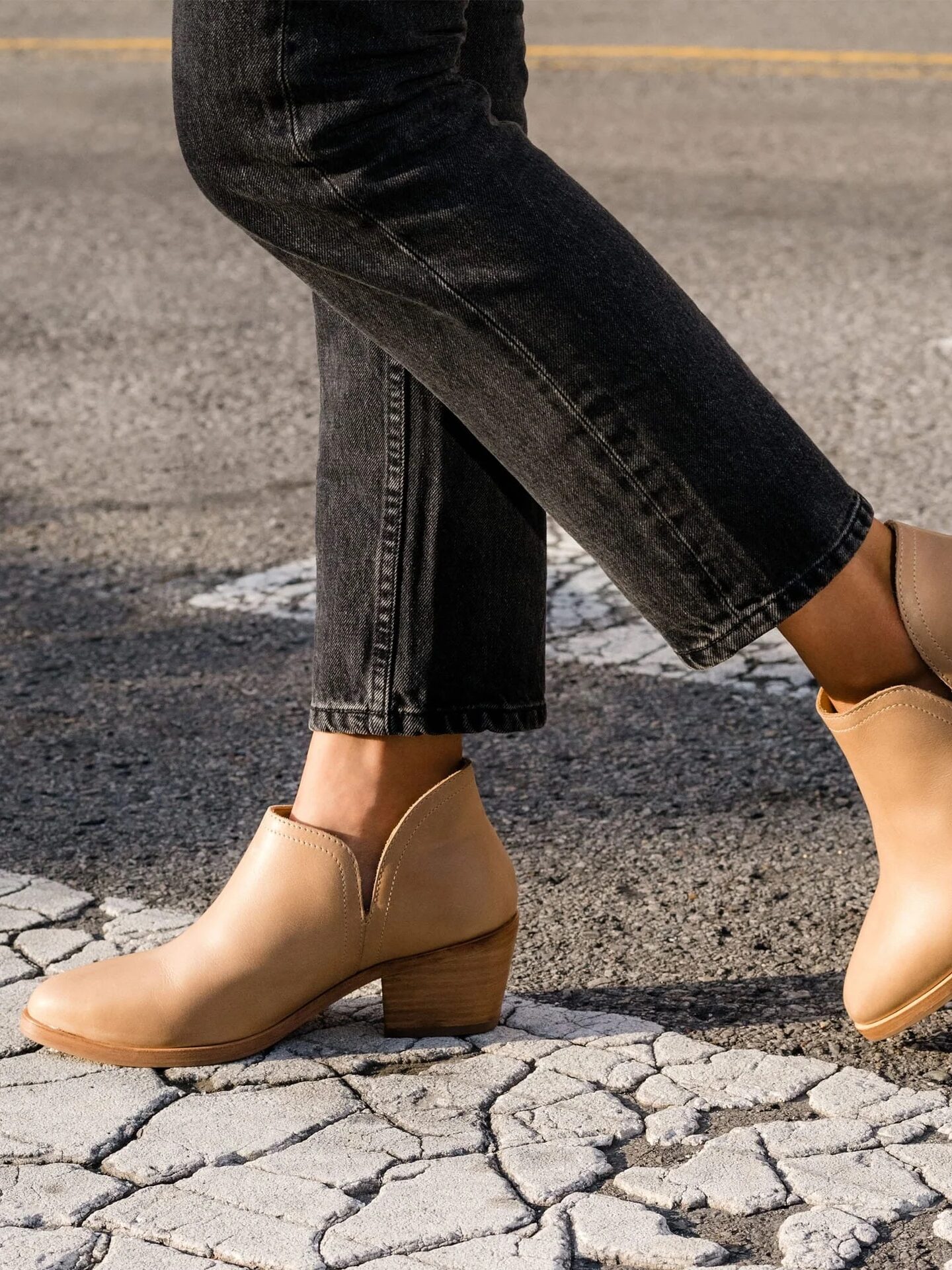 Person wearing light brown heeled ankle boots and black jeans while crossing a street with faded white paint lines.