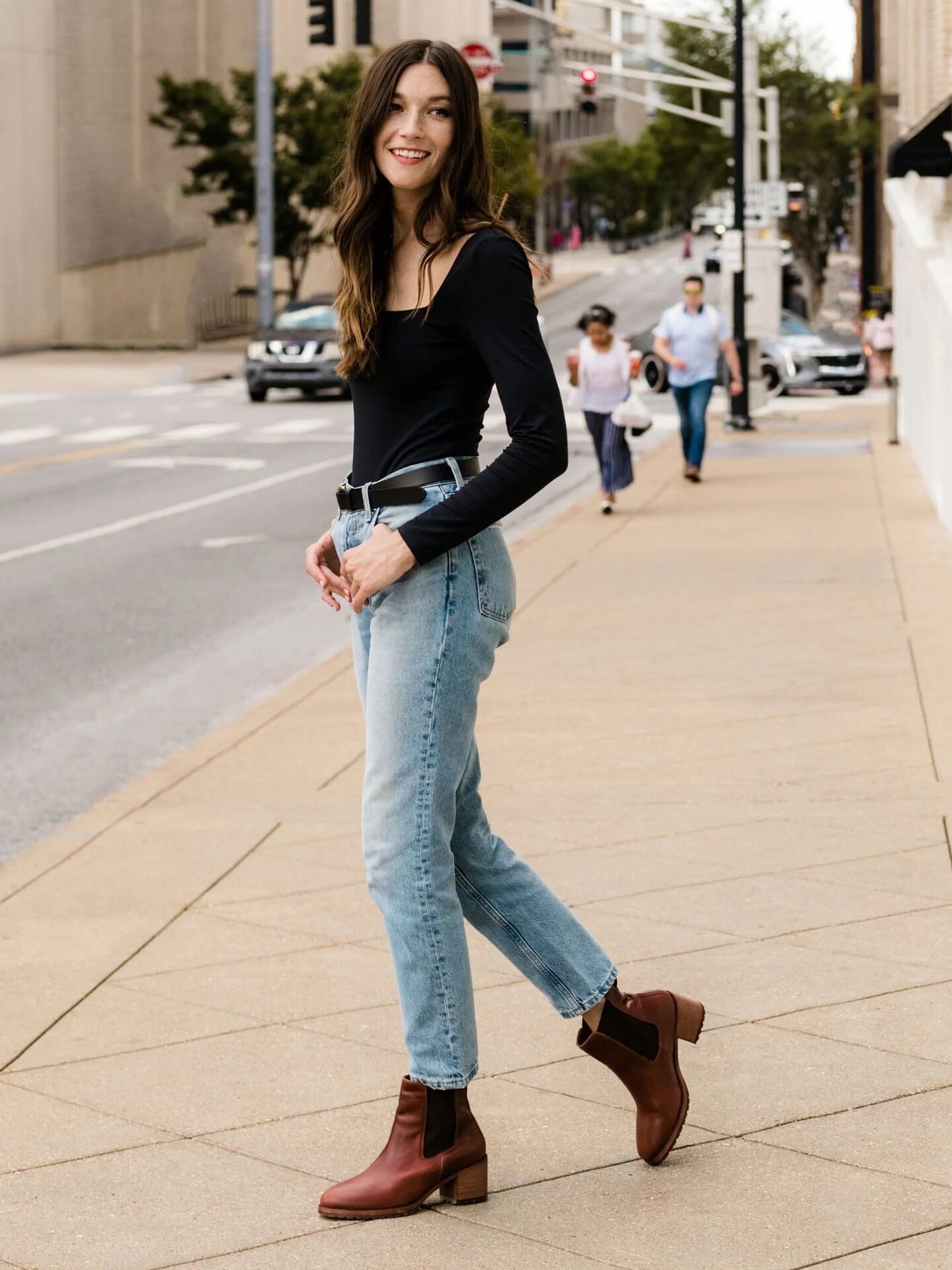 A woman in a black top and blue jeans walks on a city sidewalk, smiling and looking over her shoulder. She is wearing brown ankle boots. In the background, there are pedestrians and cars.