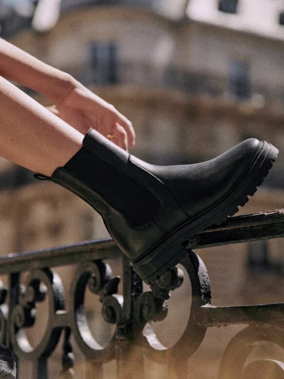 Close-up of a person wearing black ankle boots while standing on a wrought iron balcony railing with a blurred building in the background.