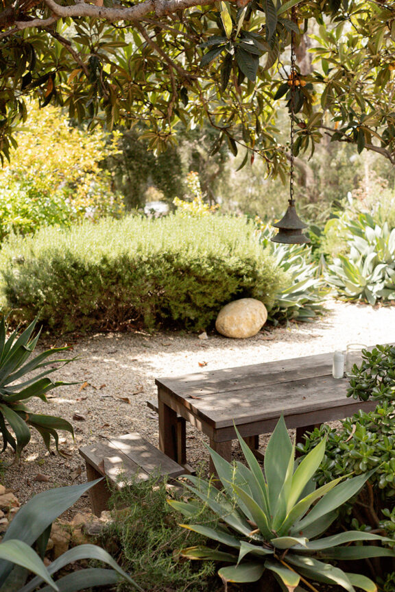 A rustic wooden picnic table sits under a tree in a garden with lush green plants and shrubs. A hanging pendant light is above the table.