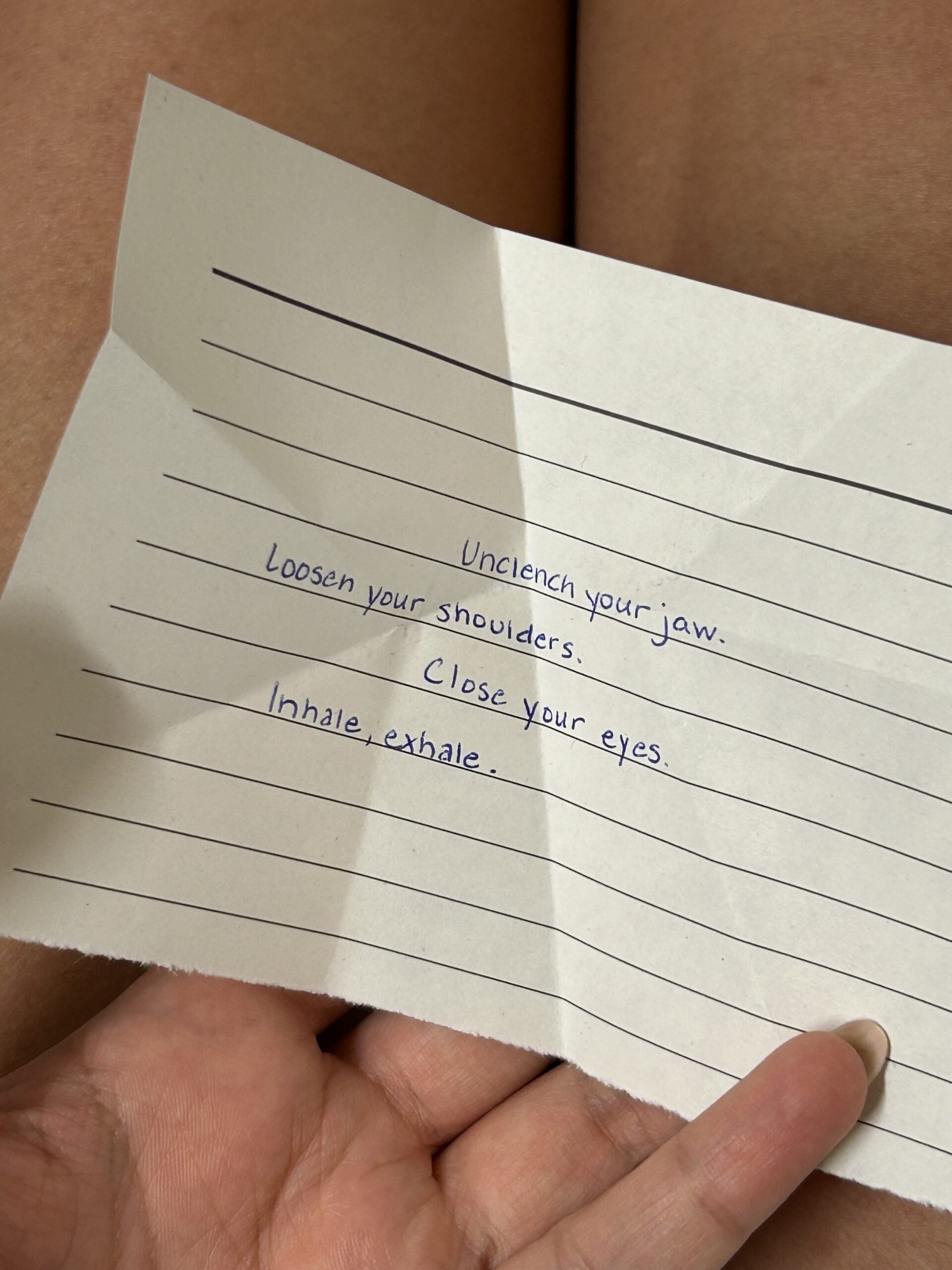 A handwritten note on lined paper says: "Unclench your jaw. Loosen your shoulders. Close your eyes. Inhale, exhale." A person holds the paper.