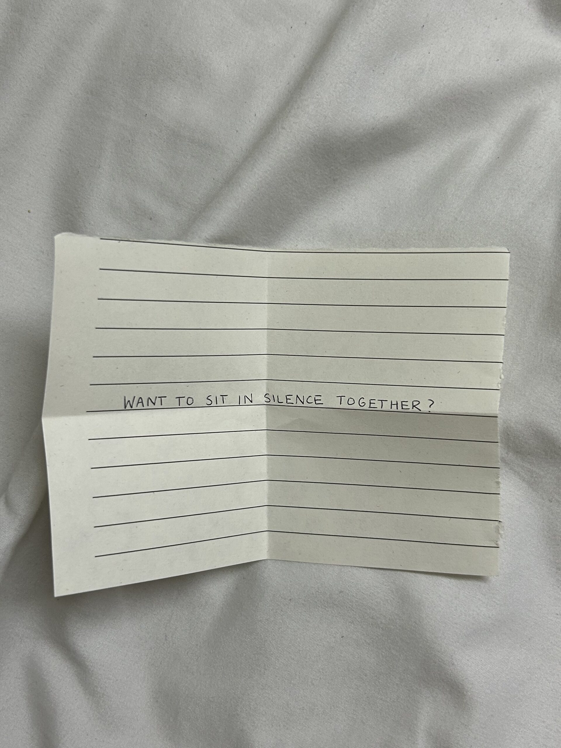 A small piece of lined paper with the handwritten text: "WANT TO SIT IN SILENCE TOGETHER?" against a soft, light-colored fabric background.