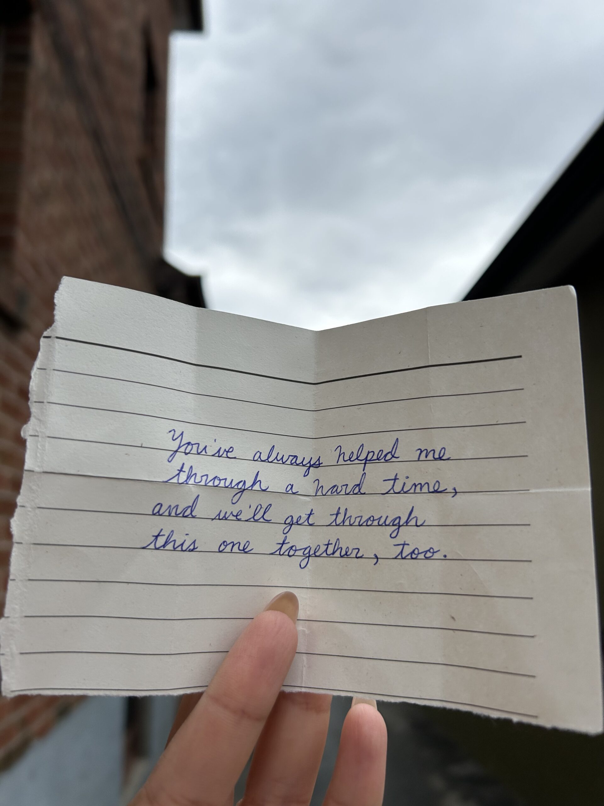 A hand holds a piece of torn, lined paper with a handwritten note that reads: "You've always helped me through a hard time, and we'll get through this one together, too.