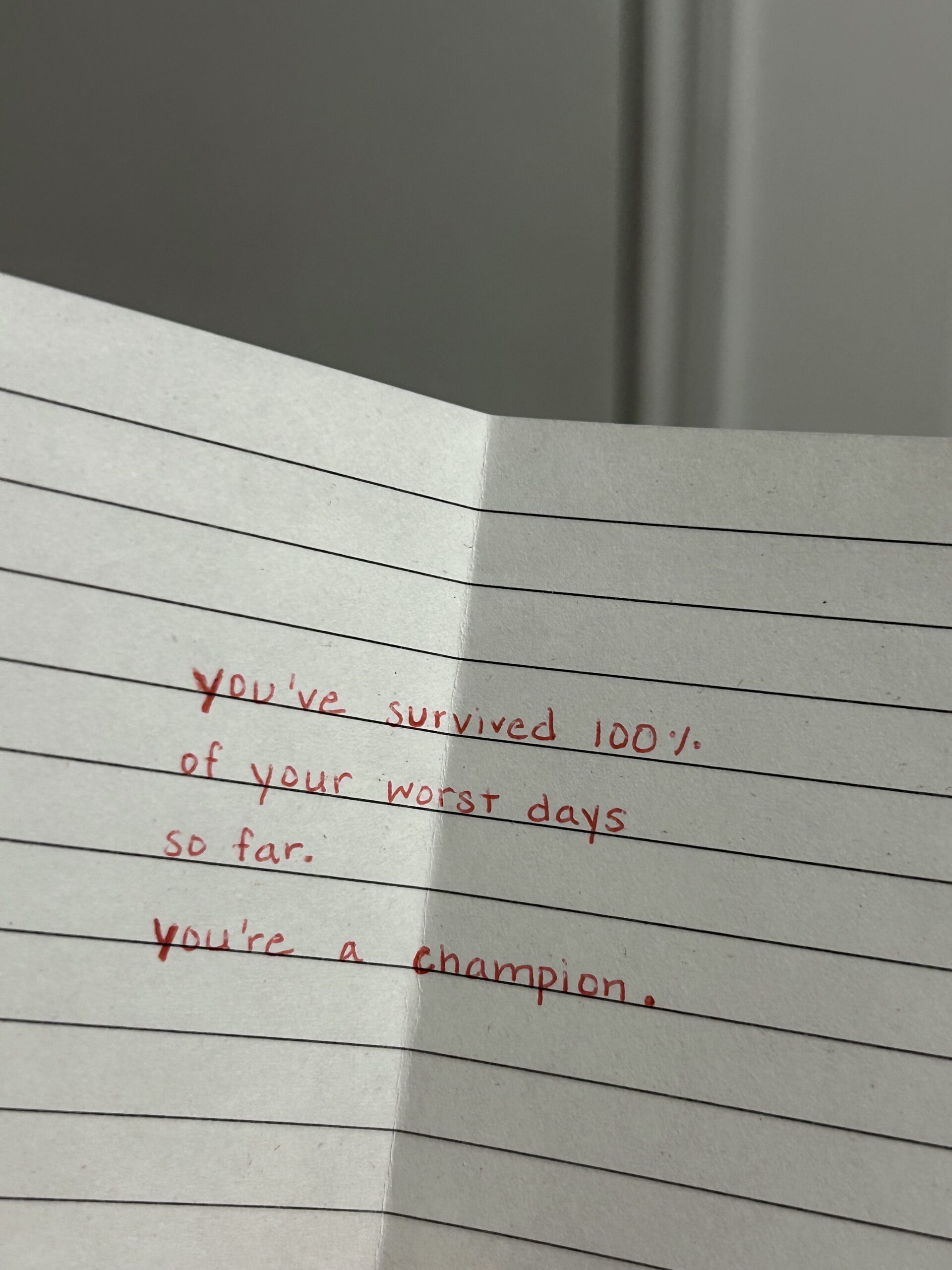 A piece of lined notebook paper with a handwritten message in red ink that reads: "You’ve survived 100% of your worst days so far. You’re a champion.