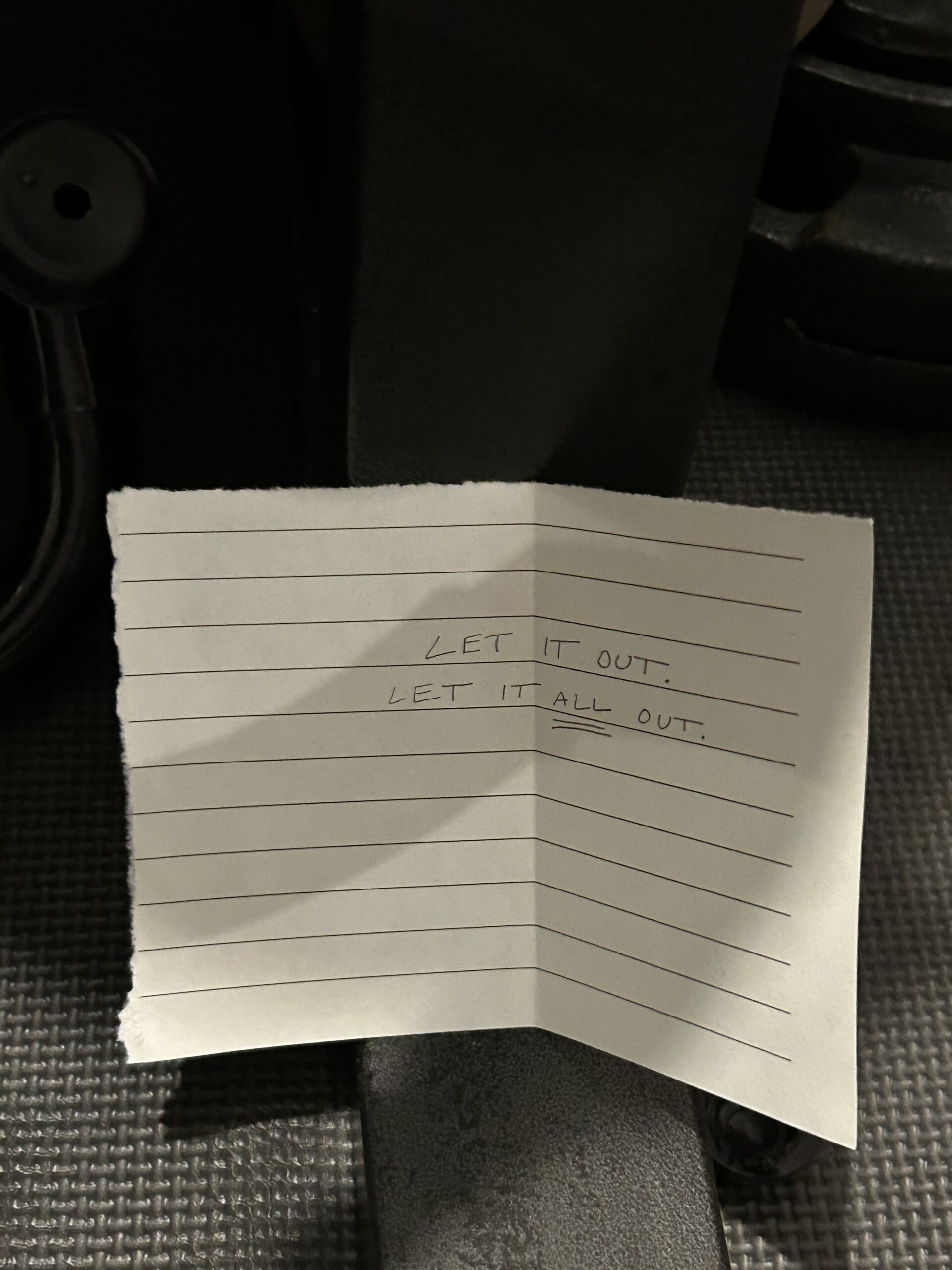 A hand-written note on lined paper reads "LET IT OUT. LET IT ALL OUT." The note is resting on a dark textured surface.