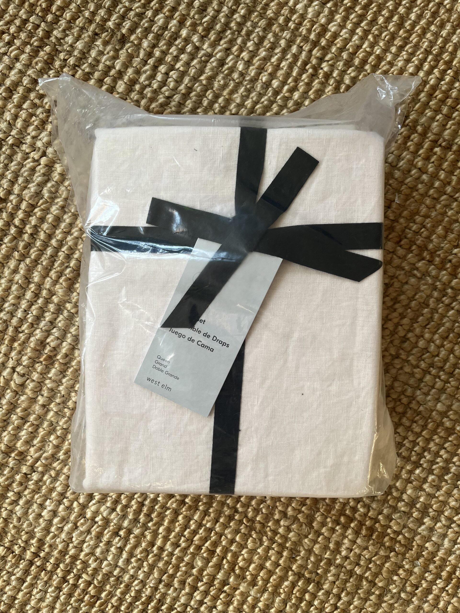 A rectangular white package wrapped in plastic with a black ribbon, placed on a textured woven mat. A label with text is attached to the ribbon.