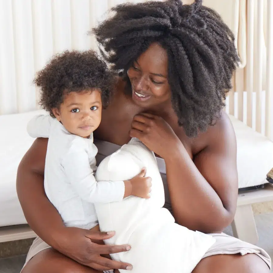 A woman with curly hair sitting on a bed, holding a small child dressed in a white outfit. The woman is smiling at the child who is looking at the camera.