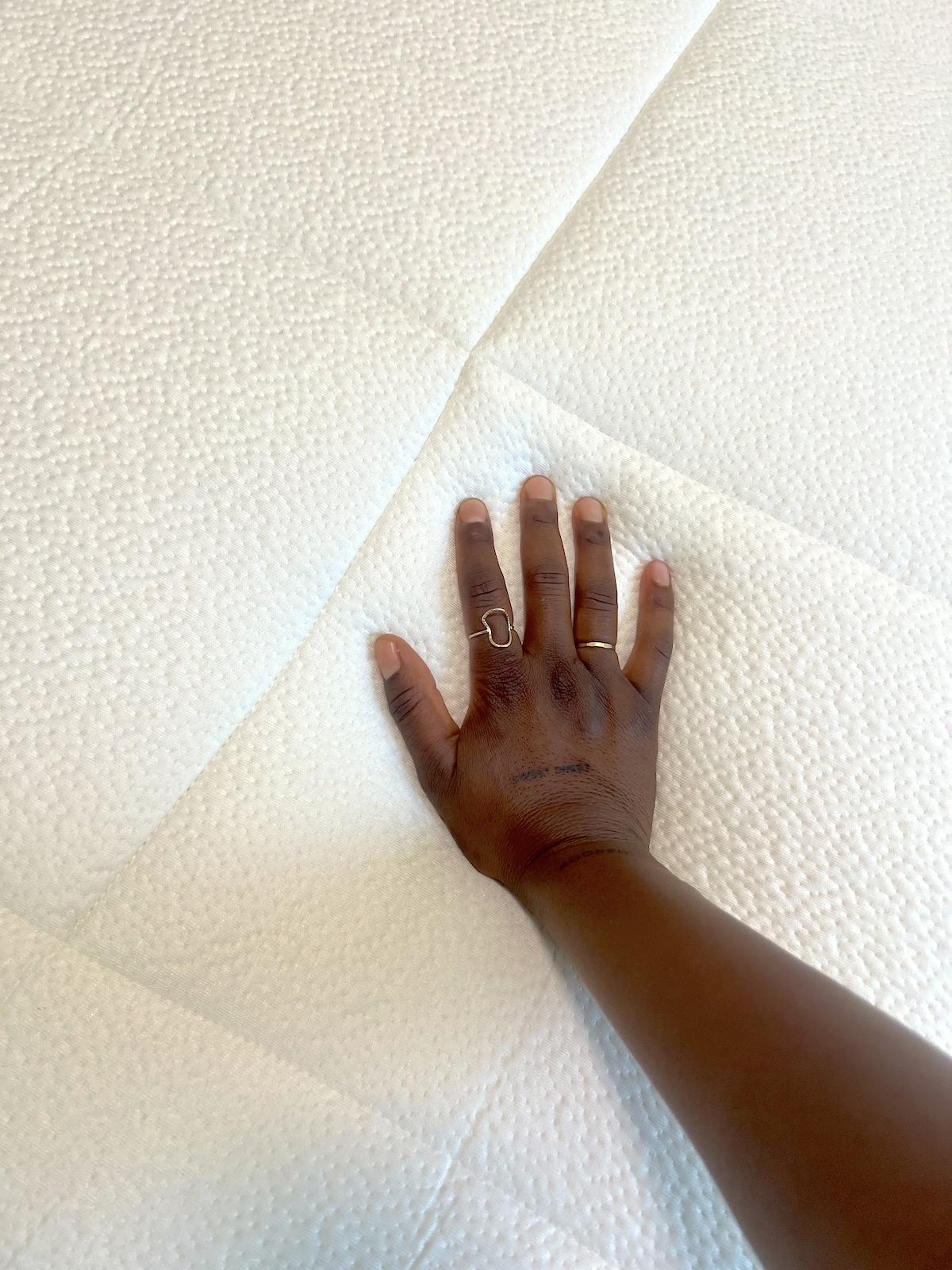 A hand with rings presses down on a white, textured surface, revealing the gentle indentation of fingers on an organic mattress.