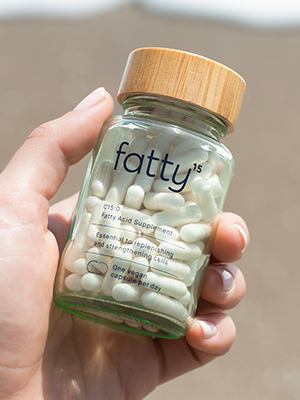 A hand holds a glass bottle labeled "fatty15," containing white capsules, against a blurred outdoor background.