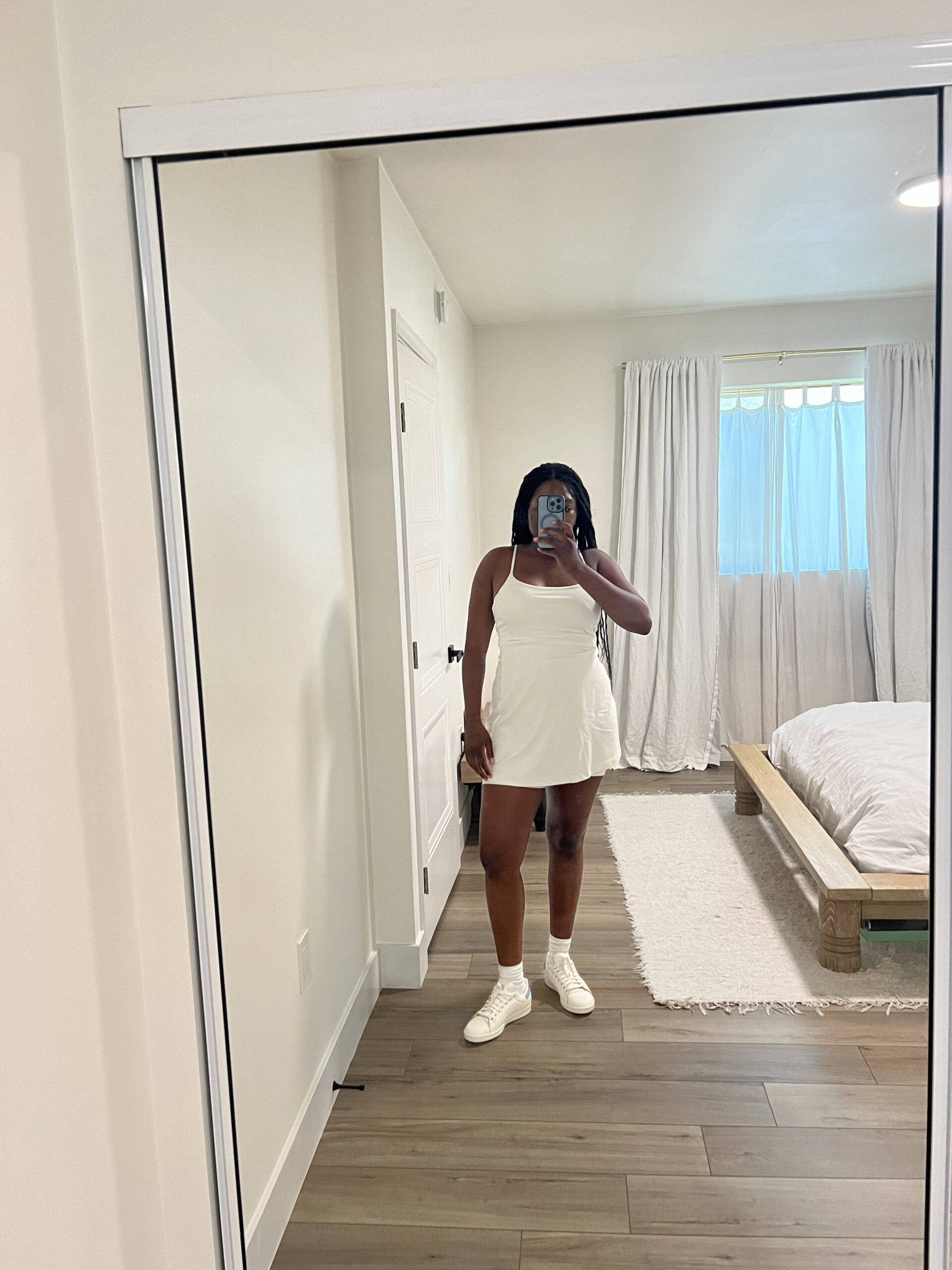 Person taking a mirror selfie in a bedroom while wearing a white dress and white shoes. The room has white walls, a bed with white bedding, and a large window with white curtains.