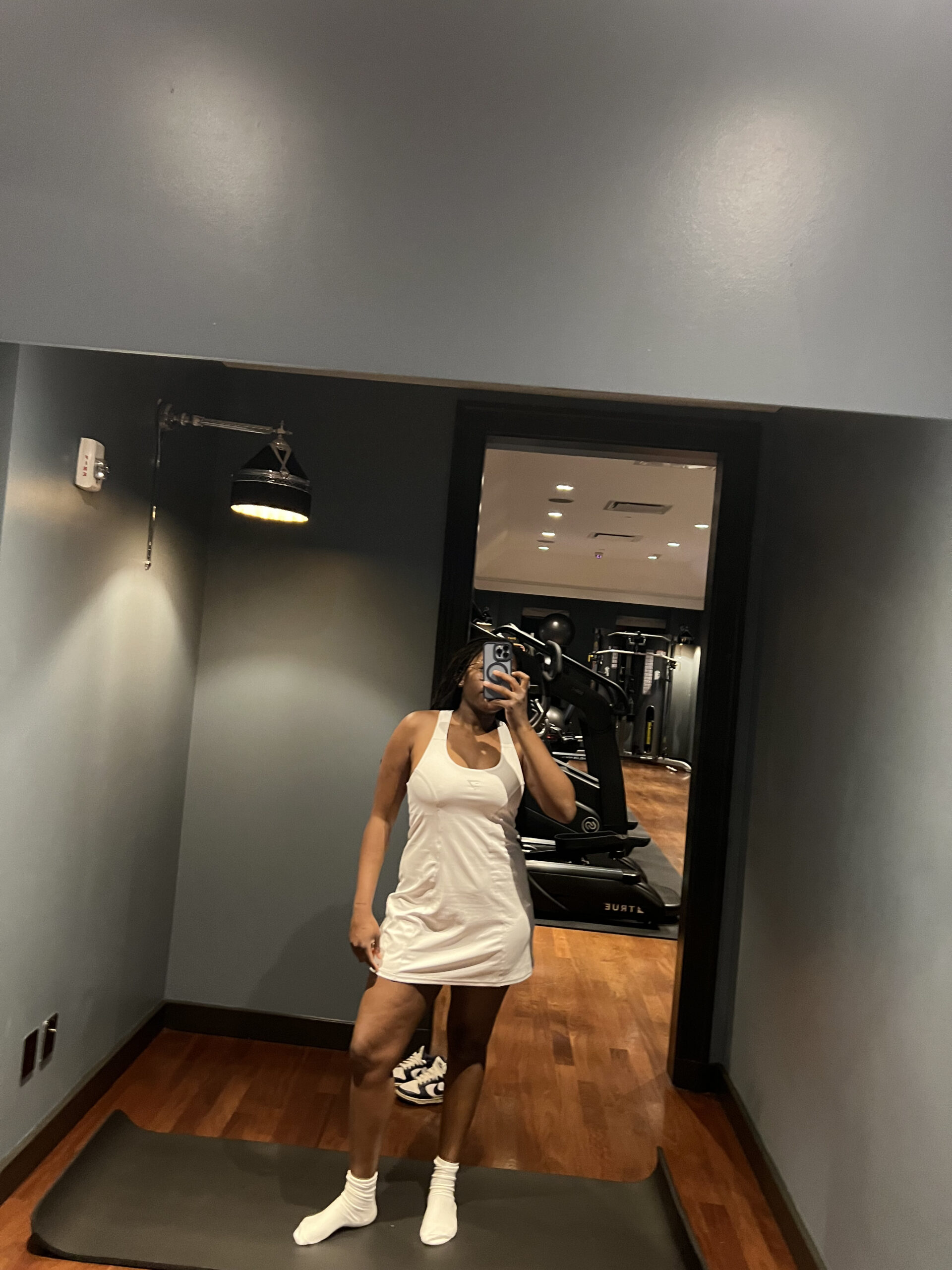 A person in a white athletic outfit takes a mirror selfie in a dimly lit gym, standing on a mat with gym equipment visible in the background.