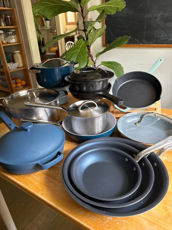 A variety of cookware, including pots, pans, and skillets, arranged on a wooden table. A potted plant is in the background.