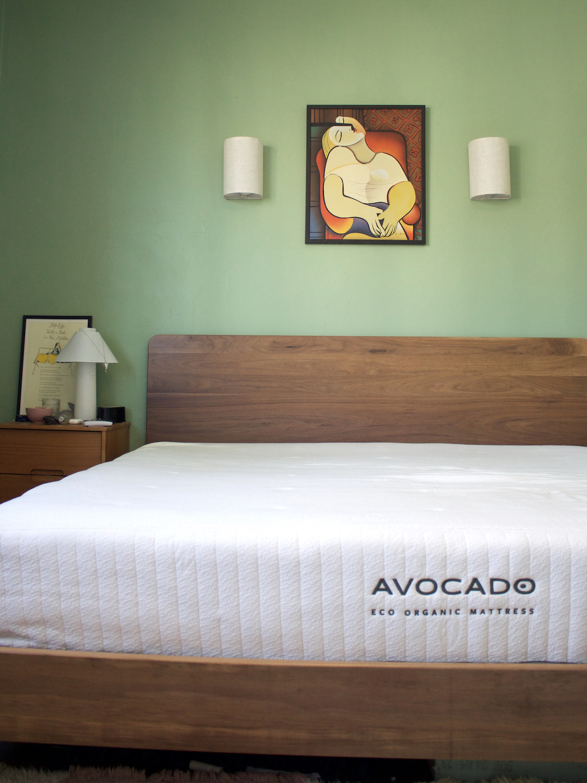 A bedroom with a wooden bed frame and an Avocado Eco Organic Mattress. The walls are painted green and there is a framed art piece above the bed, flanked by two wall-mounted lights.