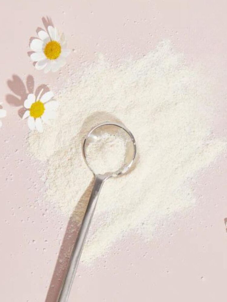 A metal spoon rests on a pile of white powder on a light pink surface, surrounded by four small white daisies with yellow centers.