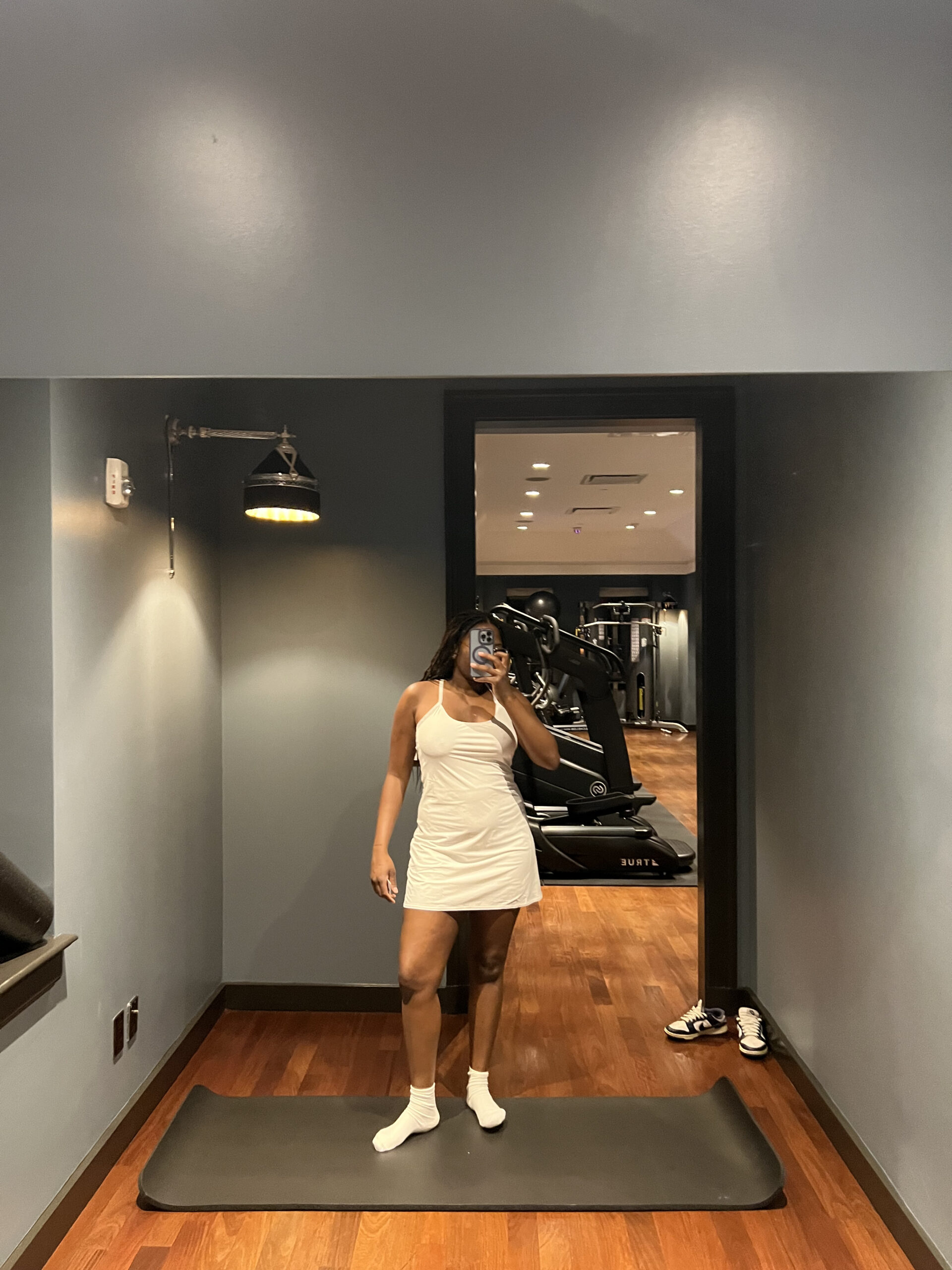 A person wearing a white dress and socks takes a selfie in a mirror. A gym with various equipment is visible in the background.