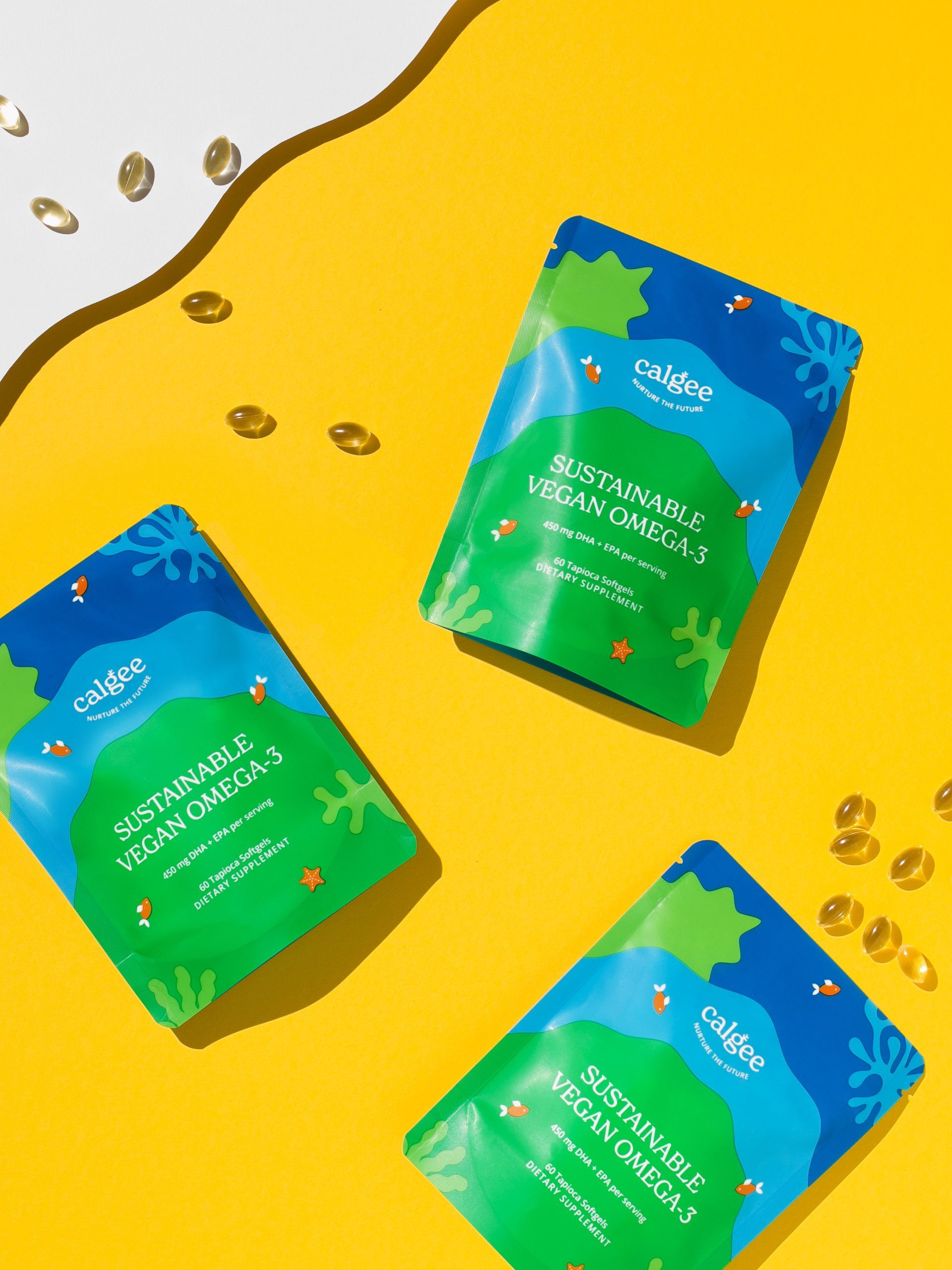 Three brightly colored packets labeled "Sustainable Vegan Omega-3" lie on a yellow surface, surrounded by scattered omega-3 capsules.