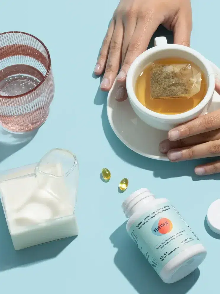 A person's hands holding a cup of tea with a tea bag. Next to them are a glass of water, a small pitcher of milk, two capsules, and an open bottle of dietary supplements on a light blue surface.