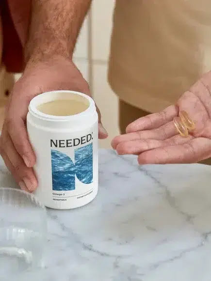 Two individuals are seen, with one holding an open container labeled "Needed" and the other holding a few capsules over a marble countertop.