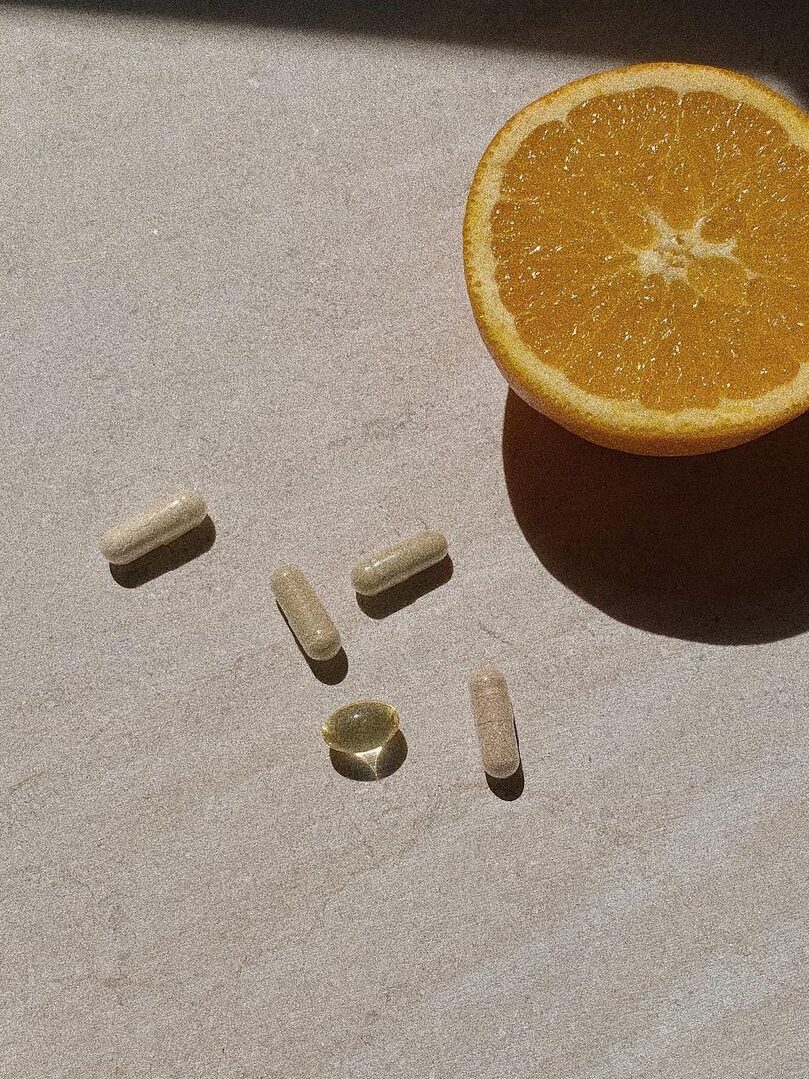 A halved orange is placed on a light surface next to five supplements, including four beige capsules and one yellow softgel.