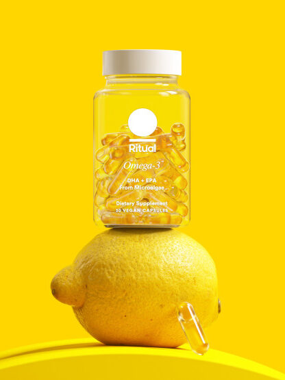 A clear jar of omega-3 capsules with the brand "Ritual" stands on a lemon. One capsule is placed next to the lemon. The background is solid yellow.