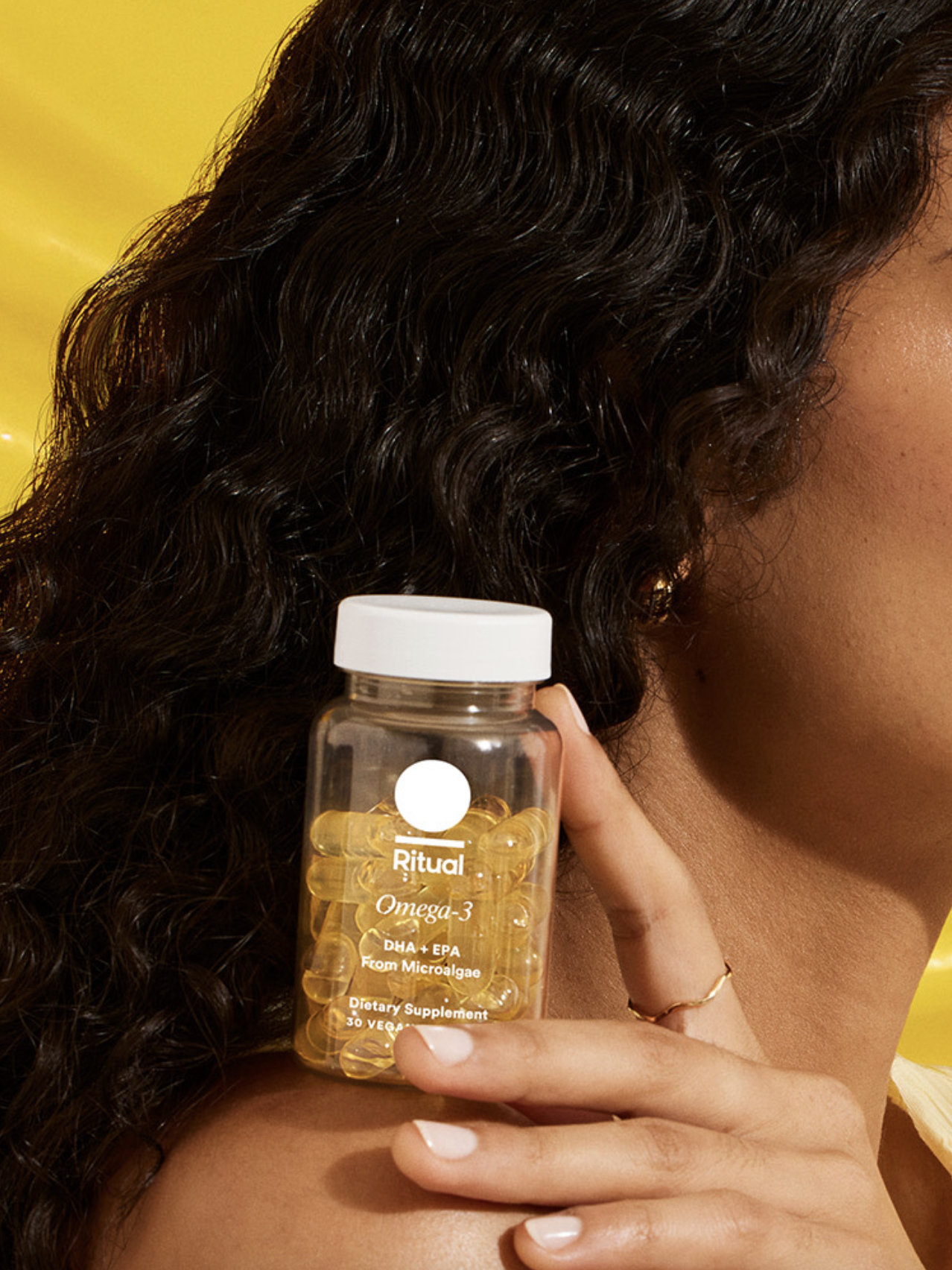 Person holding a clear bottle of Ritual Omega-3 dietary supplement capsules against a yellow background.