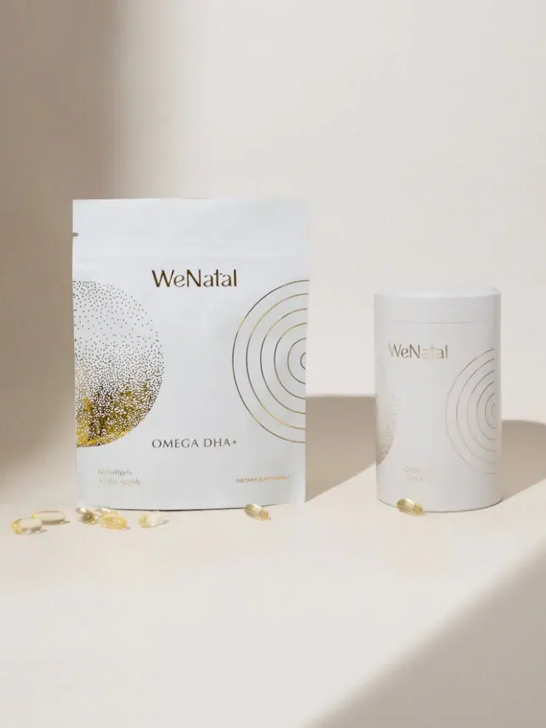 A white pouch and a white cylindrical container labeled "WeNatal Omega DHA+" are positioned on a light background, with several omega-3 softgel capsules scattered around them.