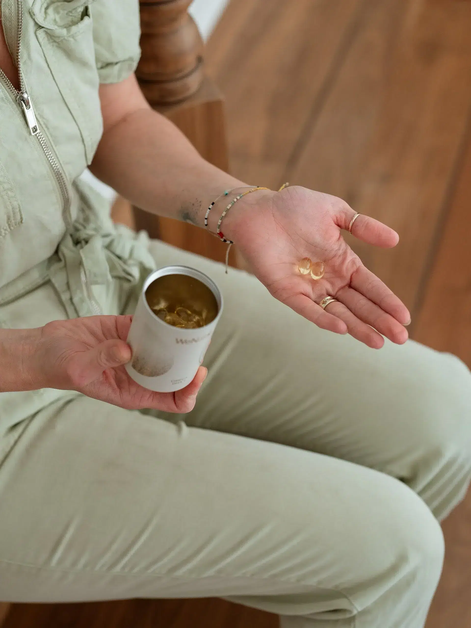 A person wearing a light green outfit holds an open container of supplements in one hand and displays several capsules in the other hand, seated on a wooden surface.