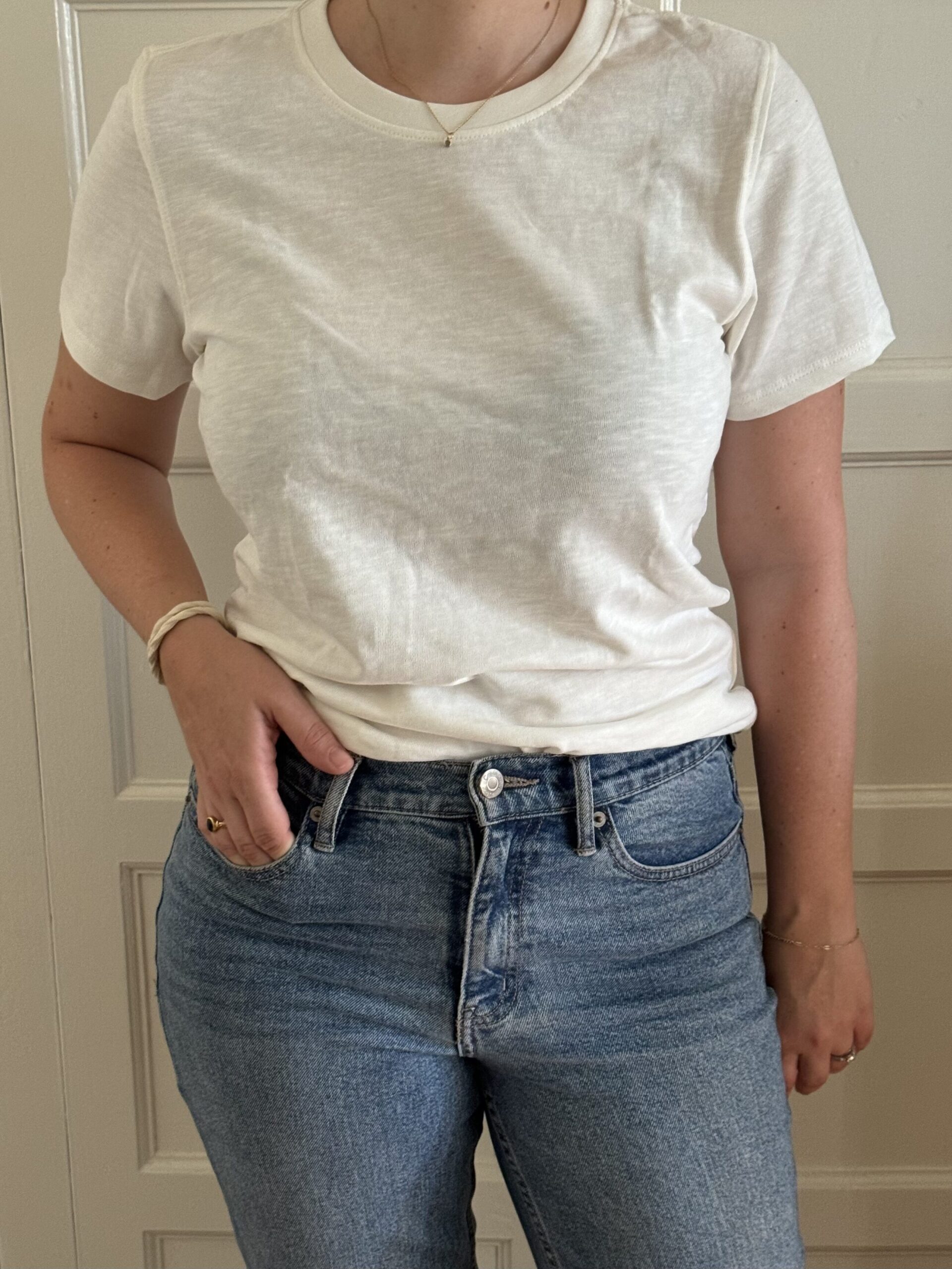 Person standing in front of a door, wearing a plain white t-shirt and blue jeans, with one hand in their pocket.