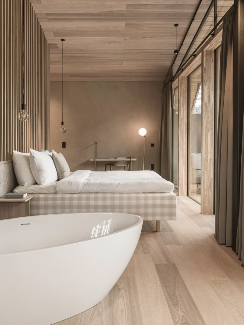 A modern, minimalist bedroom with a freestanding bathtub next to large windows offering a scenic outdoor view. The room features wooden accents, a bed, pendant lights, and a small desk.