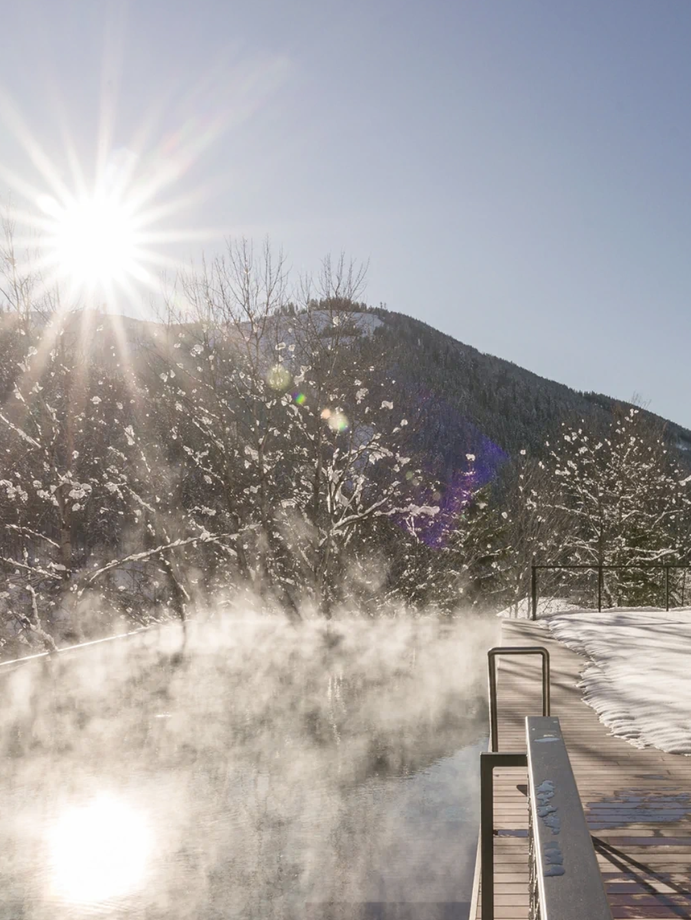 A modern wooden building stands beside a steaming outdoor pool on a sunny winter day, with snow-covered terrain and a mountain in the background.