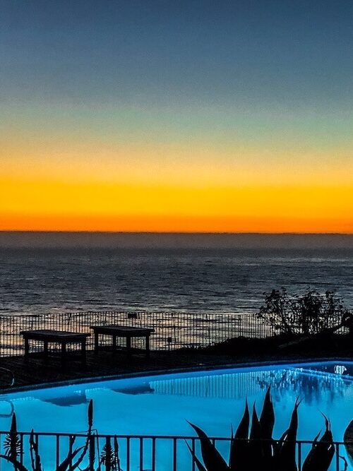 A swimming pool overlooks a tranquil ocean during sunset, with benches and plants silhouetted in the foreground.