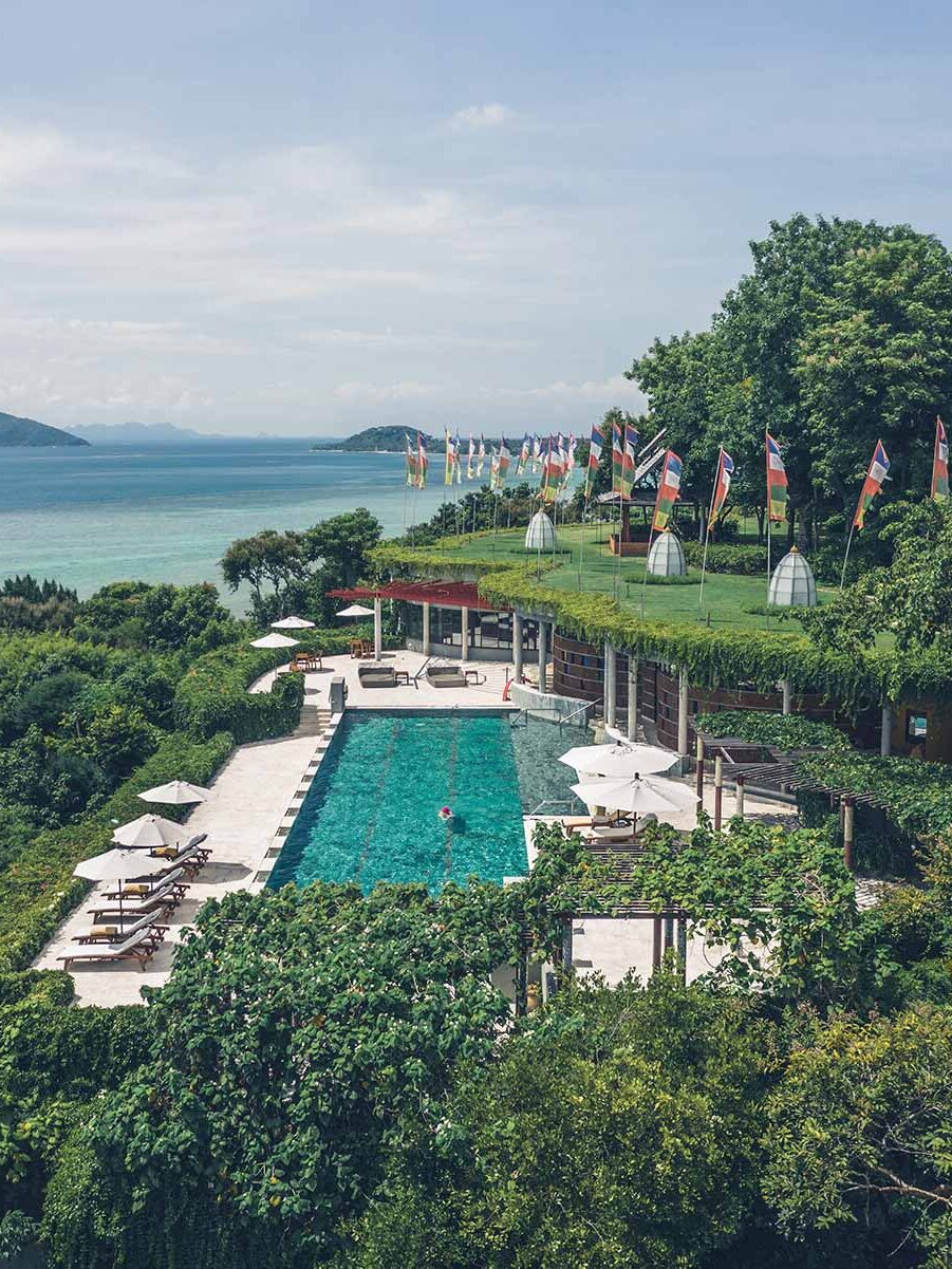 Aerial view of a lush resort featuring an infinity pool surrounded by greenery. Numerous flags line the edge of the pool area. The sea and distant islands are visible in the background.