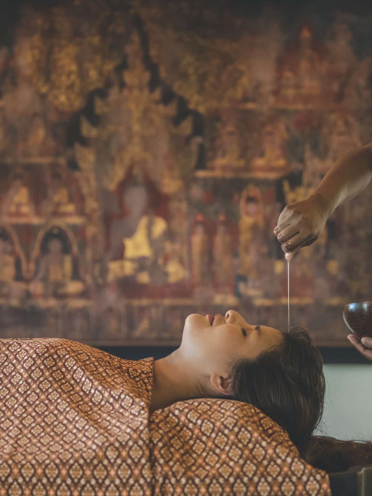 A person lies on a table receiving an oil treatment on their forehead from a practitioner in a yellow uniform, with an intricate wall painting in the background.