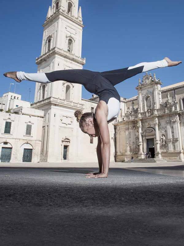 A person performs a handstand split in front of an Italian baroque-style church with a tall bell tower on a sunny day.