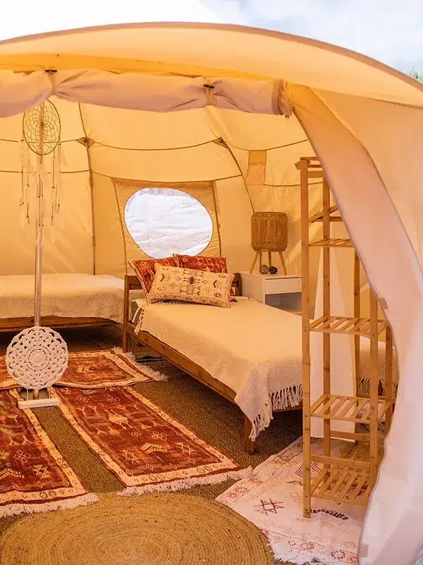 A cozy interior of a glamping tent with two single beds, a wooden shelf, dreamcatchers, and patterned rugs on the floor.