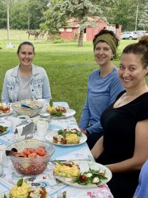 A group of eight women sit around a picnic table outdoors, enjoying a meal together. The table is set with various dishes, including eggs, bacon, and fruit. Trees and a red barn are visible in the background.